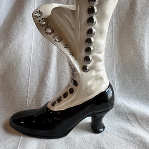 May include: A ceramic boot-shaped vase in black and white. The boot features a glossy black foot and heel, with a white upper section adorned with silver-colored buttons and decorative cutouts. The vase is designed to resemble a vintage boot.