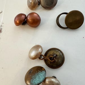 May include: A collection of vintage cufflinks in various shapes and finishes. The cufflinks include round, oval, and button-style designs in metallic tones of silver, bronze, and copper. Some have a tarnished, aged appearance.
