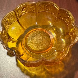 May include: A vintage amber-colored glass bowl with a scalloped edge and floral detailing. The bowl has two small handles and a circular base. The glass has a textured appearance, and the bowl is likely used for serving or display.