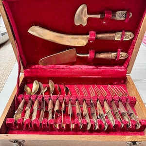 May include: A vintage wooden cutlery set with a red velvet interior. The set includes brass-coloured serving utensils and a collection of knives, forks, and spoons with wooden handles. The case is open, revealing the neatly arranged silverware.