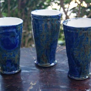 May include: Three handmade ceramic vases with a deep blue glaze and a white interior. The vases have a textured surface with brown and green accents. The vases are of varying heights and sit on a dark surface.