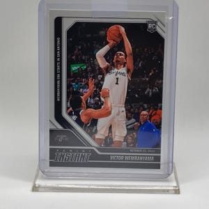 Op de afbeelding: Een basketbal trading card met Victor Wembanyama in een Spurs-uniform, halverwege een sprong, een basketbal schietend. De kaart zit in een doorzichtige beschermhoes. Tekst bevat "WEMBANYAMA ERA STARTS IN SAN ANTONIO" en "INSTANT".