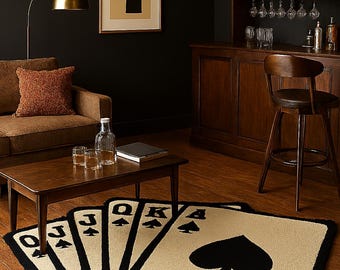 May include: A decorative rug shaped like a hand of playing cards, featuring the Ace, King, Queen, Jack, and Ten of Spades. The rug has a beige background with black borders and card symbols. The rug is placed on a wooden floor.