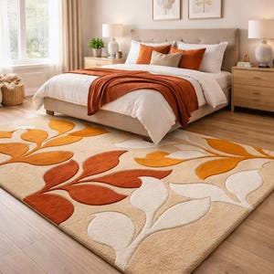 May include: A bedroom scene featuring a beige rug with a leaf design in orange and white. The rug is in front of a bed with white linens and orange pillows and throw. The room has light-colored walls and wooden furniture.