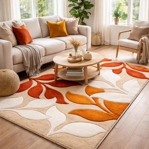 May include: Beige area rug with a leaf design in shades of orange and white. The rug is placed in a living room with a white sofa, wooden coffee table, and a chair. The room has large windows and natural light.