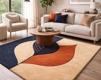 May include: A large area rug with a leaf design in shades of navy blue, orange, and beige. The rug is placed in a living room with a sofa, chairs, and a wooden coffee table. The rug's dimensions are not visible.