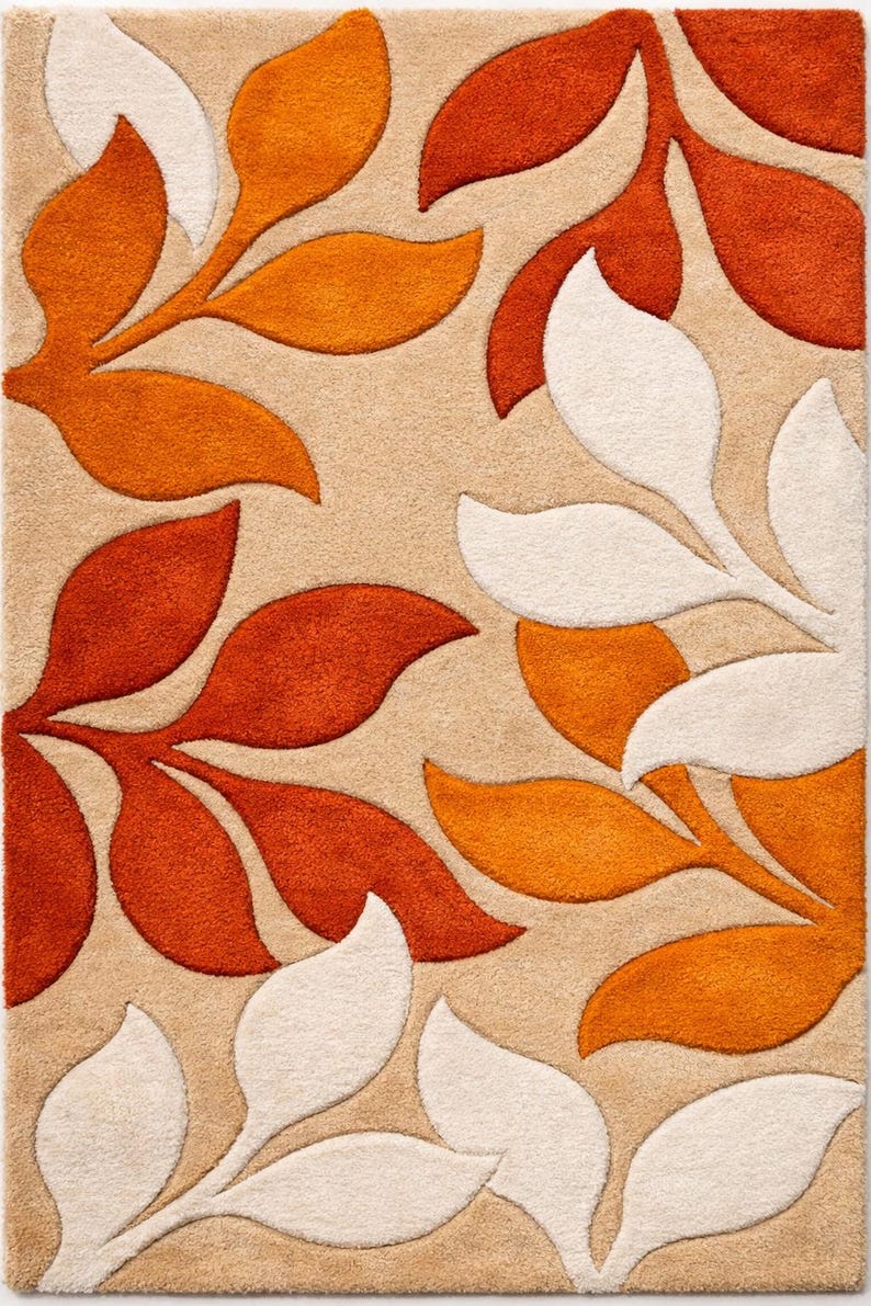 May include: A beige area rug with a repeating leaf pattern in shades of orange and white. The leaves are stylized and arranged across the rug's surface, creating a textured, decorative design. The rug is suitable for living rooms and bedrooms.