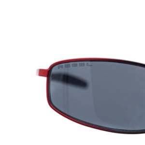 May include: Red-framed sunglasses with dark gray lenses. The word "REBEL" is printed on the lens. The sunglasses have a modern, rectangular shape and are set against a white background.