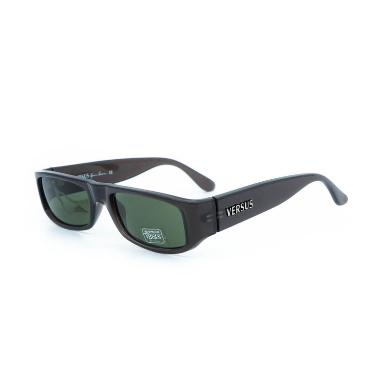 K&ouml;nnte beinhalten: Dunkelbraune Sonnenbrille mit rechteckigem Rahmen und gr&uuml;nen Gl&auml;sern. Der Markenname "VERSUS" ist wei&szlig; auf dem B&uuml;gel aufgedruckt. Die Sonnenbrille ist vor einem wei&szlig;en Hintergrund platziert.
