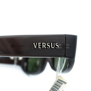 K&ouml;nnte beinhalten: Nahaufnahme einer dunkelbraunen Sonnenbrille mit dem Wort &bdquo;VERSUS&ldquo; in silbernen Buchstaben auf dem B&uuml;gel. Die Gl&auml;ser sind gr&uuml;n, und ein wei&szlig;es Etikett h&auml;ngt am Rahmen. Die Sonnenbrille ist vor wei&szlig;em Hintergrund.