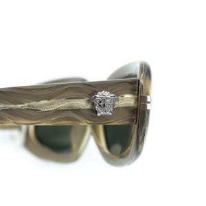 May include: Close-up of gold-toned sunglasses with a wavy design on the frames. A silver-coloured Medusa head emblem is on the temple. The lenses are a dark green colour. The sunglasses are set against a white background.