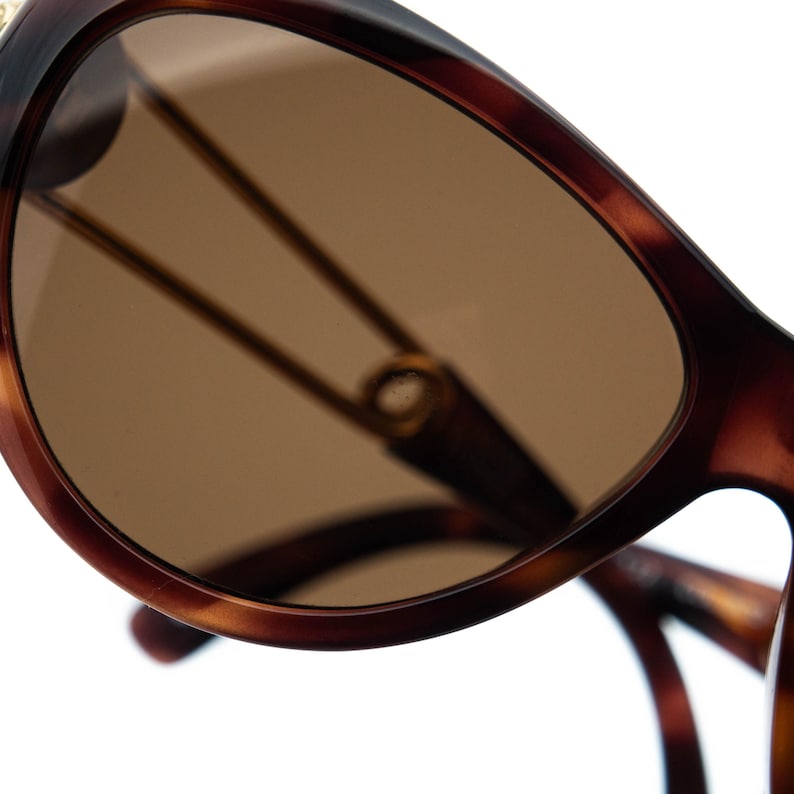 K&ouml;nnte beinhalten: Nahaufnahme einer Schildpatt-Sonnenbrille mit braunen Gl&auml;sern. Der Rahmen hat eine klassische, abgerundete Form mit einem dunkelbraunen und bernsteinfarbenen Muster. Die Gl&auml;ser sind warmbraun, und die Metallb&uuml;gel sind sichtbar.
