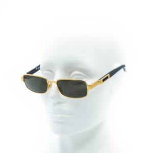 May include: Gold-framed sunglasses with rectangular dark lenses. The sunglasses have black temples and a gold bridge. The brand name is visible on the temple. The sunglasses are displayed on a white mannequin head.