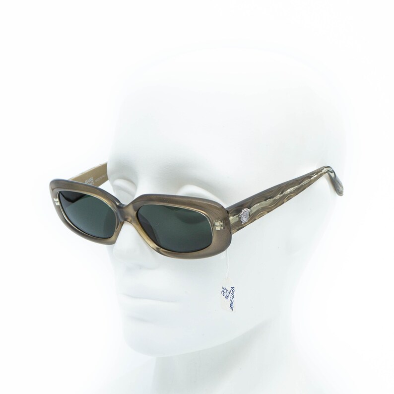 May include: Light brown rectangular sunglasses with dark lenses. The temples feature a textured pattern and a silver emblem near the hinges. The sunglasses are displayed on a white mannequin head.