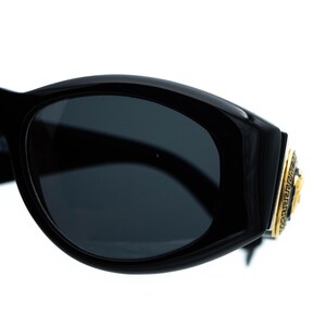 May include: Black sunglasses with dark lenses. The frames are thick and glossy, with a gold-colored emblem on the temple featuring a Medusa head and a Greek key design. The sunglasses have a bold, rectangular shape.