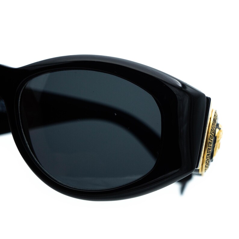 May include: Black sunglasses with dark lenses. The frames are thick and glossy, with a gold-colored emblem on the temple featuring a Medusa head and a Greek key design. The sunglasses have a bold, rectangular shape.
