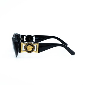 May include: Black sunglasses with a rectangular gold frame and a Medusa head emblem. The sunglasses have thick, curved temples and a glossy finish. The brand name is visible on the temple.