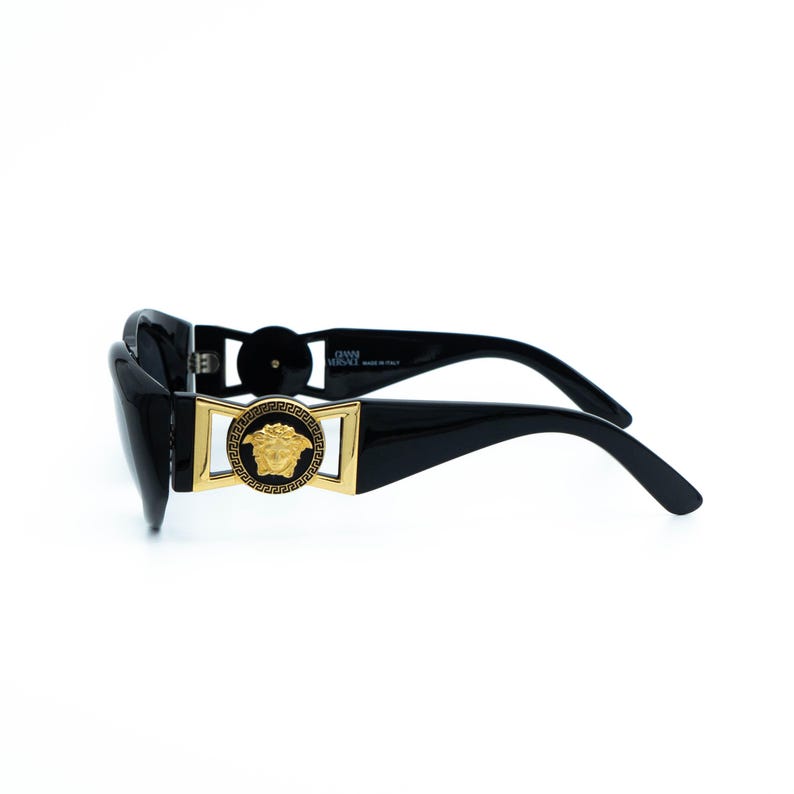 May include: Black sunglasses with a rectangular gold frame and a Medusa head emblem. The sunglasses have thick, curved temples and a glossy finish. The brand name is visible on the temple.