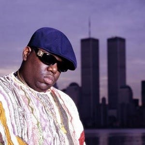 May include: Portrait of a man wearing a navy blue cap, sunglasses, and a patterned shirt. A gold chain is visible around his neck. The background features the Twin Towers and a purple sky.