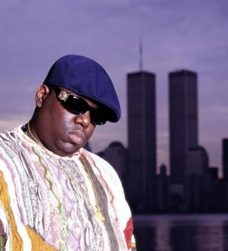 May include: Portrait of a man wearing a navy blue cap, sunglasses, and a patterned shirt. A gold chain is visible around his neck. The background features the Twin Towers and a purple sky.
