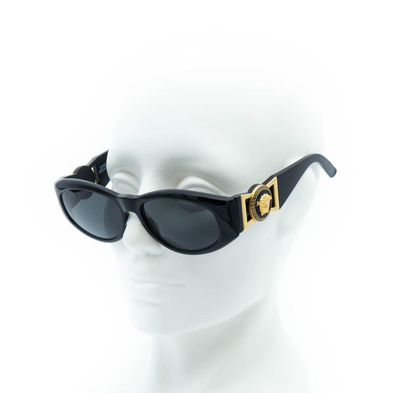 May include: Black oval sunglasses with dark lenses. The sunglasses feature gold-colored accents and a Medusa head emblem on the temples. The frames are glossy and appear to be made of a durable material.