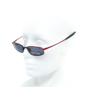 May include: Red and black sunglasses with dark lenses. The sunglasses have a rectangular shape and the text "REBEL" is visible on the lens. The arms of the sunglasses are red with black tips. The sunglasses are displayed on a white mannequin head.