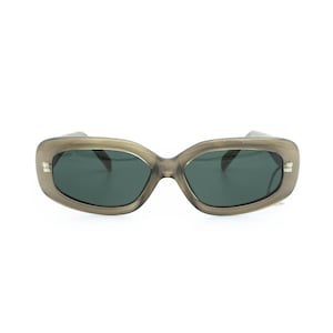 May include: Oval sunglasses with a translucent, taupe-coloured frame and dark lenses. The sunglasses have a rectangular shape and small silver accents on the temples. The lenses are a dark green colour.