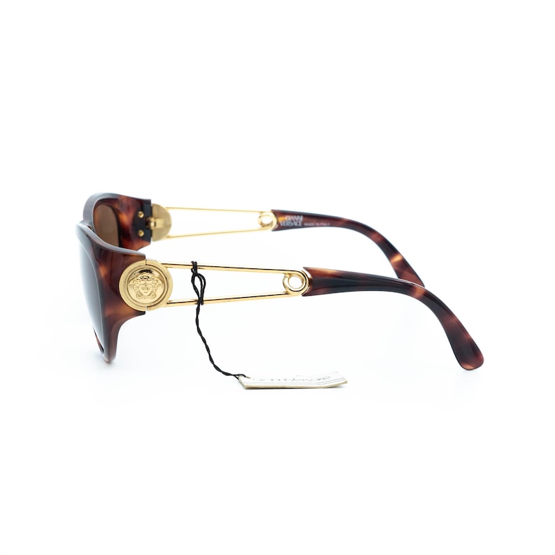 K&ouml;nnte beinhalten: Sonnenbrille in Schildpatt-Optik mit goldfarbenen Akzenten. Die Sonnenbrille hat ein Medusa-Kopf-Emblem auf dem Rahmen und ein Sicherheitsnadel-Design an den B&uuml;geln. Die Gl&auml;ser sind braun get&ouml;nt. Ein kleines Etikett h&auml;ngt am Rahmen.