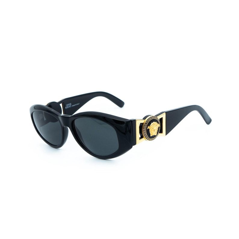 May include: Black oval sunglasses with dark lenses. The sunglasses feature a gold-colored Medusa head emblem on the temples. The frames are glossy and the arms curve slightly downward.