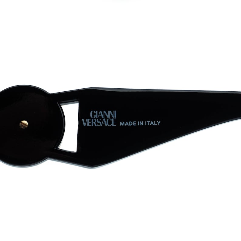 May include: Black sunglasses with a unique geometric design. The text "GIANNI VERSACE MADE IN ITALY" is printed on the frame. A small gold-colored accent is visible on the frame.