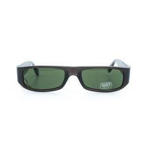 K&ouml;nnte beinhalten: Dunkelgraue Sonnenbrille mit rechteckigen gr&uuml;nen Gl&auml;sern. Die Sonnenbrille hat einen dicken Rahmen und einen kleinen Aufkleber auf dem rechten Glas mit der Aufschrift "100% PROTECTION VERSUS".