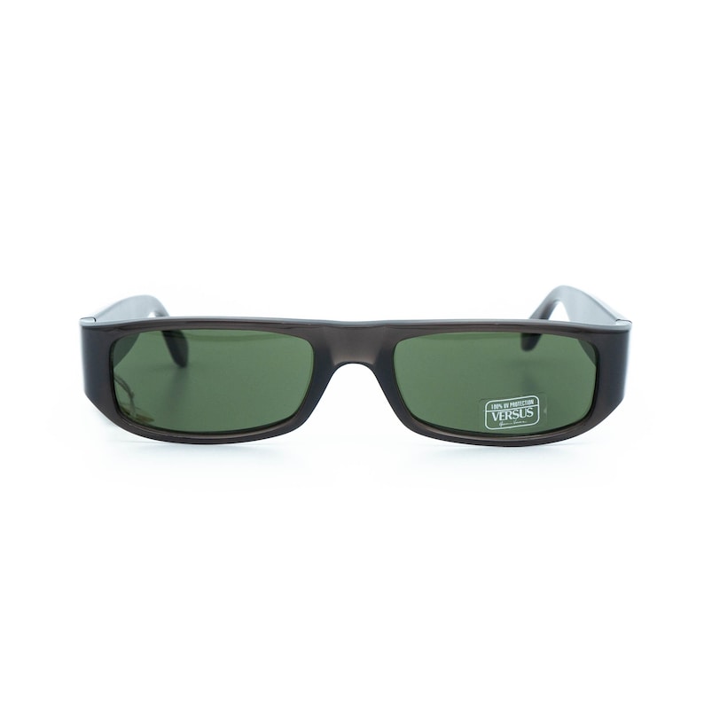 K&ouml;nnte beinhalten: Dunkelgraue Sonnenbrille mit rechteckigen gr&uuml;nen Gl&auml;sern. Die Sonnenbrille hat einen dicken Rahmen und einen kleinen Aufkleber auf dem rechten Glas mit der Aufschrift "100% PROTECTION VERSUS".