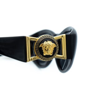 May include: Black sunglasses with a gold Medusa head emblem. The sunglasses have a rectangular gold frame and a black, glossy finish. The design is a classic, luxury style.