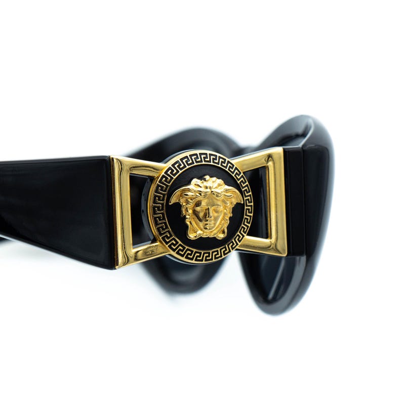 May include: Black sunglasses with a gold Medusa head emblem. The sunglasses have a rectangular gold frame and a black, glossy finish. The design is a classic, luxury style.