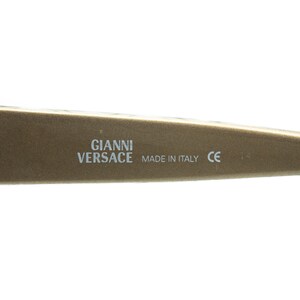 May include: Close-up of a section of a gold-coloured eyeglass frame. The text "GIANNI VERSACE MADE IN ITALY CE" is printed in white. The frame has a smooth, matte finish.