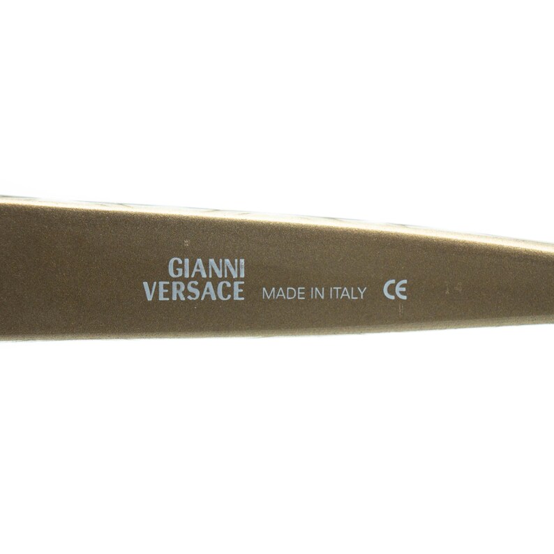 May include: Close-up of a section of a gold-coloured eyeglass frame. The text "GIANNI VERSACE MADE IN ITALY CE" is printed in white. The frame has a smooth, matte finish.