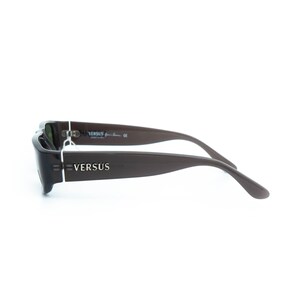 K&ouml;nnte beinhalten: Dunkelbraune Versus-Sonnenbrille mit rechteckigem Rahmen und dunklen Gl&auml;sern. Der Markenname "VERSUS" ist in silbernen Buchstaben auf dem B&uuml;gel abgebildet. Die Sonnenbrille hat ein schlankes, modernes Design.