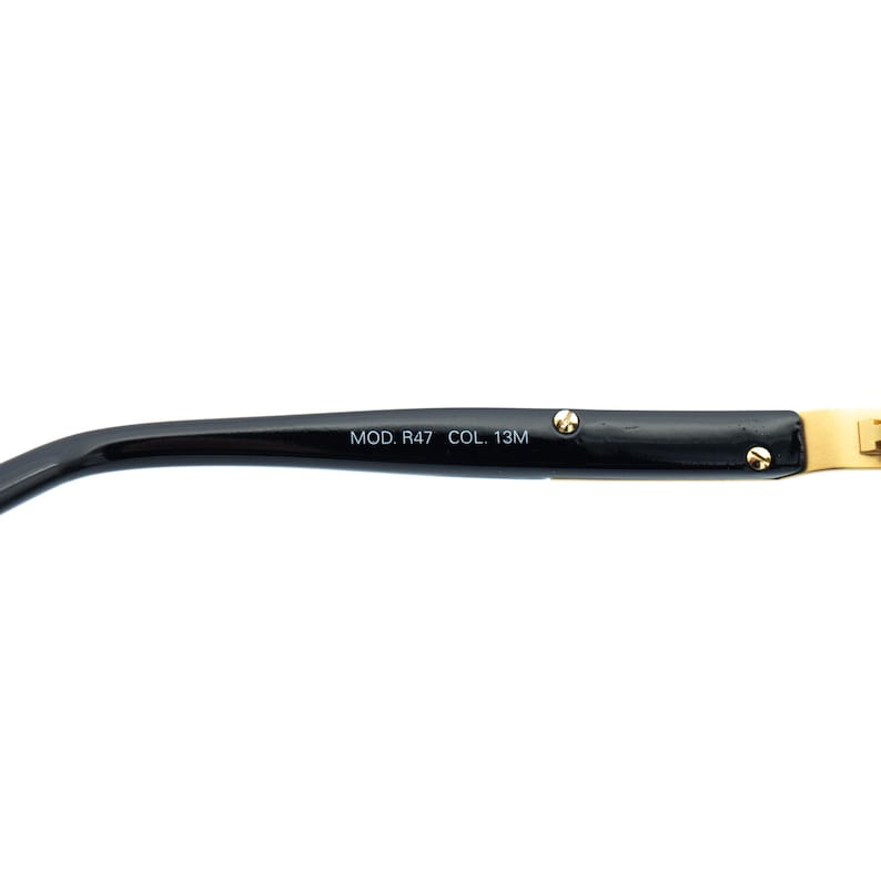 May include: Close-up of the arm of a pair of eyeglasses. The arm is black with gold-colored accents and the text "MOD. R47 COL. 13M". The glasses arm curves slightly.