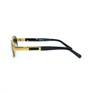May include: Gold-framed sunglasses with black temples. The sunglasses have a rectangular shape and a gold bridge. The brand name "VERSUS" is printed on the temple. The sunglasses are set against a white background.