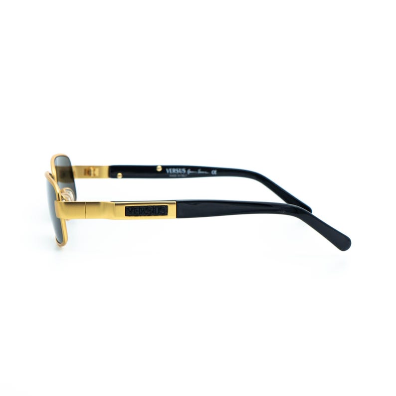 May include: Gold-framed sunglasses with black temples. The sunglasses have a rectangular shape and a gold bridge. The brand name "VERSUS" is printed on the temple. The sunglasses are set against a white background.