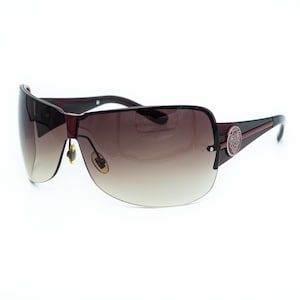 May include: Oversized shield sunglasses featuring a dark burgundy frame and a gradient brown lens. The temples are adorned with a circular emblem displaying a crest design. These sunglasses include adjustable nose pads and a sleek, wrap-around silhouette.