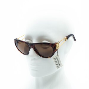 K&ouml;nnte beinhalten: Braune Sonnenbrille im Schildpatt-Muster mit goldfarbenen Akzenten. Die Sonnenbrille hat eine Cat-Eye-Form und braune Gl&auml;ser. Die B&uuml;gel sind mit einem goldfarbenen Nadeldesign mit Medusenhaupt-Emblem versehen. Ein Etikett mit dem Markennamen "Gianni Versace" ist angebracht.