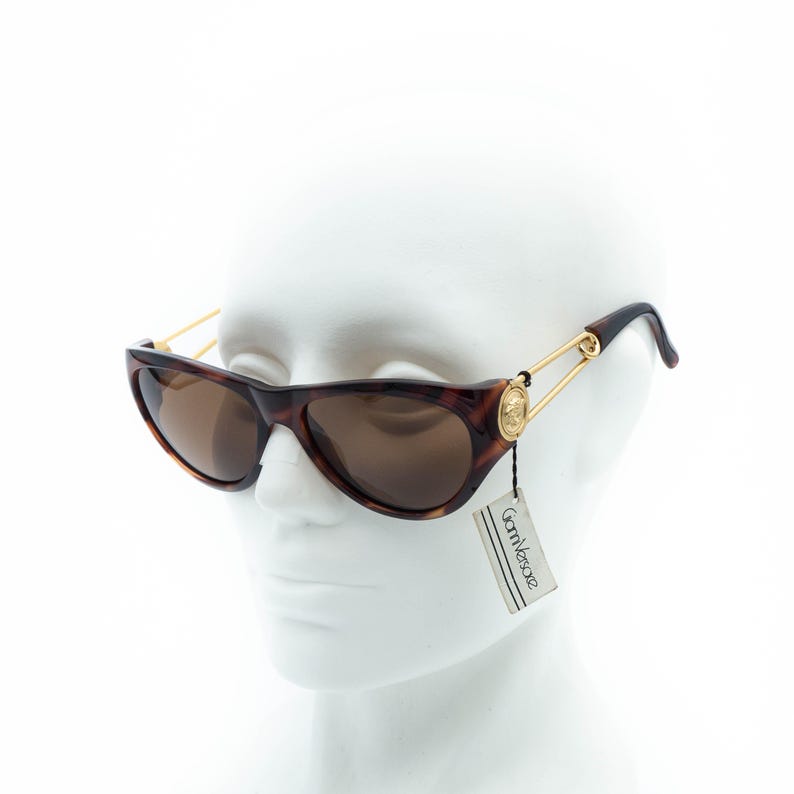 K&ouml;nnte beinhalten: Braune Sonnenbrille im Schildpatt-Muster mit goldfarbenen Akzenten. Die Sonnenbrille hat eine Cat-Eye-Form und braune Gl&auml;ser. Die B&uuml;gel sind mit einem goldfarbenen Nadeldesign mit Medusenhaupt-Emblem versehen. Ein Etikett mit dem Markennamen "Gianni Versace" ist angebracht.