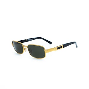 May include: Gold-framed sunglasses with dark lenses. The rectangular frames have a gold finish, with black temples and the brand name "VERSUS" printed on the side. The sunglasses are set against a white background.