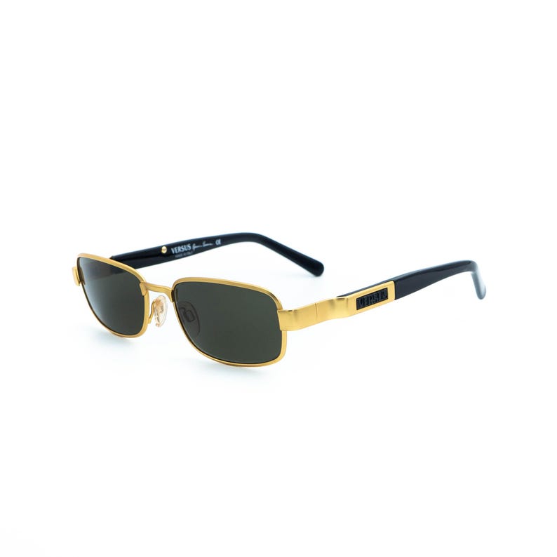 May include: Gold-framed sunglasses with dark lenses. The rectangular frames have a gold finish, with black temples and the brand name "VERSUS" printed on the side. The sunglasses are set against a white background.