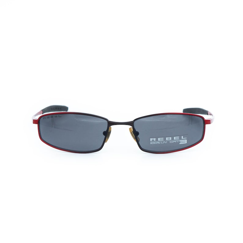 May include: Rectangular sunglasses with a black frame and red accents. The lenses are dark gray, and the text "REBEL 100% UV CAT. 3" is printed on the right lens. The sunglasses have a modern, sporty design.