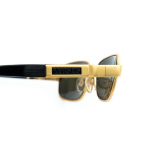 May include: Gold-toned sunglasses with black temples and dark lenses. The brand name "VERSUS" is displayed on the frame. The sunglasses have a rectangular shape and a modern design.