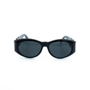 May include: Black sunglasses with a thick, rounded frame and dark lenses. The sunglasses have a sleek, modern design, suitable for various face shapes. The temples feature gold-colored accents.