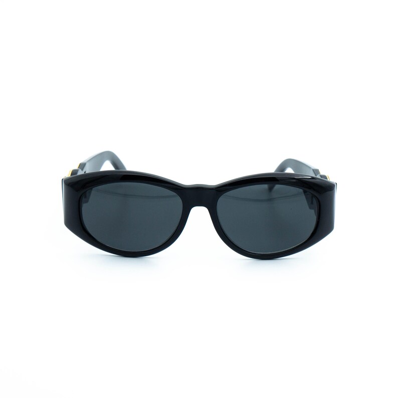 May include: Black sunglasses with a thick, rounded frame and dark lenses. The sunglasses have a sleek, modern design, suitable for various face shapes. The temples feature gold-colored accents.
