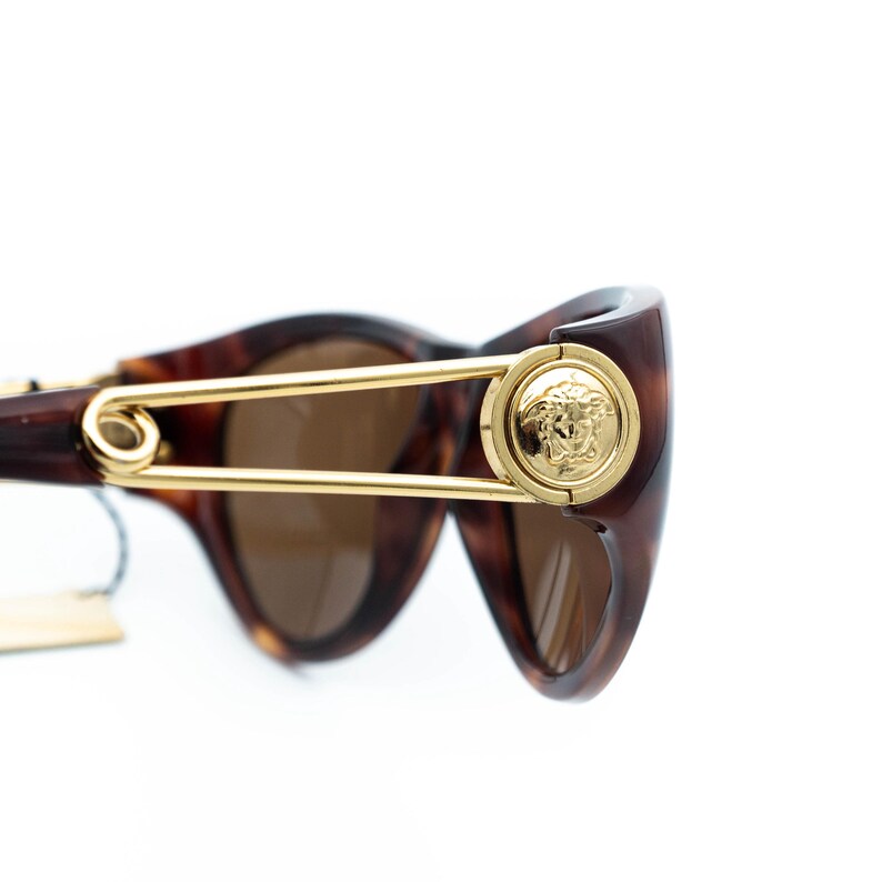 K&ouml;nnte beinhalten: Braune Sonnenbrille mit Schildpattmuster und goldfarbenen Akzenten. Die Sonnenbrille hat ein goldfarbenes Sicherheitsnadel-Design und ein goldenes Medusa-Kopf-Emblem. Die Gl&auml;ser sind braun get&ouml;nt.